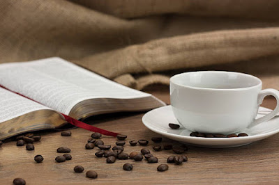 Uma xicara branca com café sobre a mesa com uma bíblia aberta e grãos de café espalhados em volta