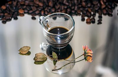 Uma xicara de café sobre um espelho, com uma rosa seca em volta e grãos de café espalhados no superior da foto
